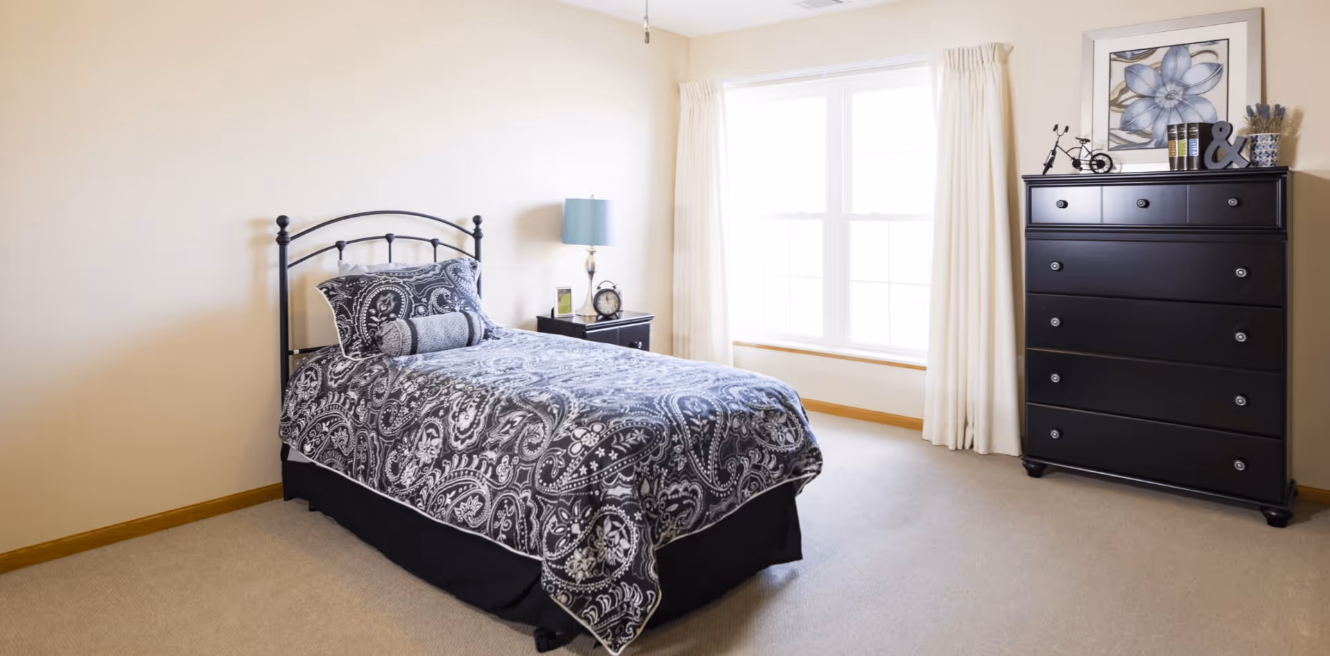 A bright bedroom with a single bed featuring a black and white patterned bedspread and matching pillows. Next to the bed is a small black nightstand with a lamp, clock, and picture frame. A large window with white curtains lets in natural light. On the right side of the room is a black dresser with decorative items and a framed flower artwork above it. The walls are light-colored and the floor is carpeted.