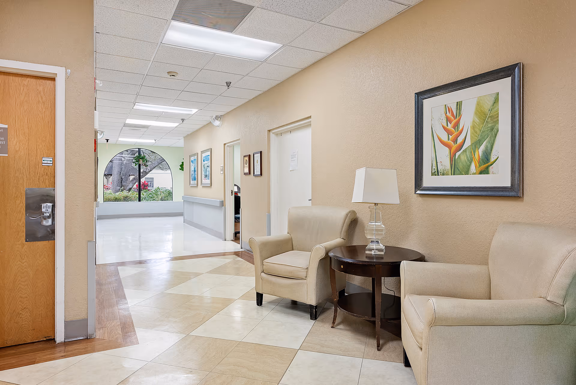Hallway seating area with two beige armchairs and a side table with a lamp, framed artwork on the wall, and a bright corridor leading to an arched window.