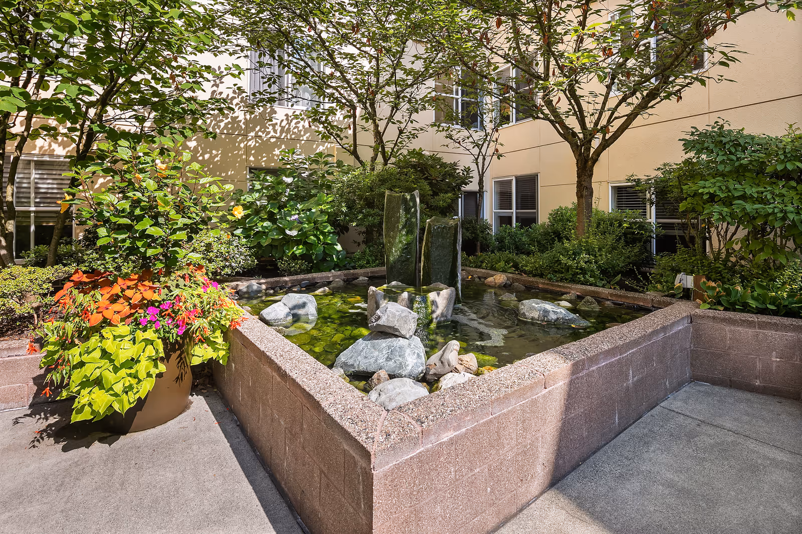 A courtyard garden area with a raised rectangular water feature containing rocks and a small fountain. Surrounding the water feature are various green plants, shrubs, and small trees. The courtyard is enclosed by the beige walls of the building with windows visible.