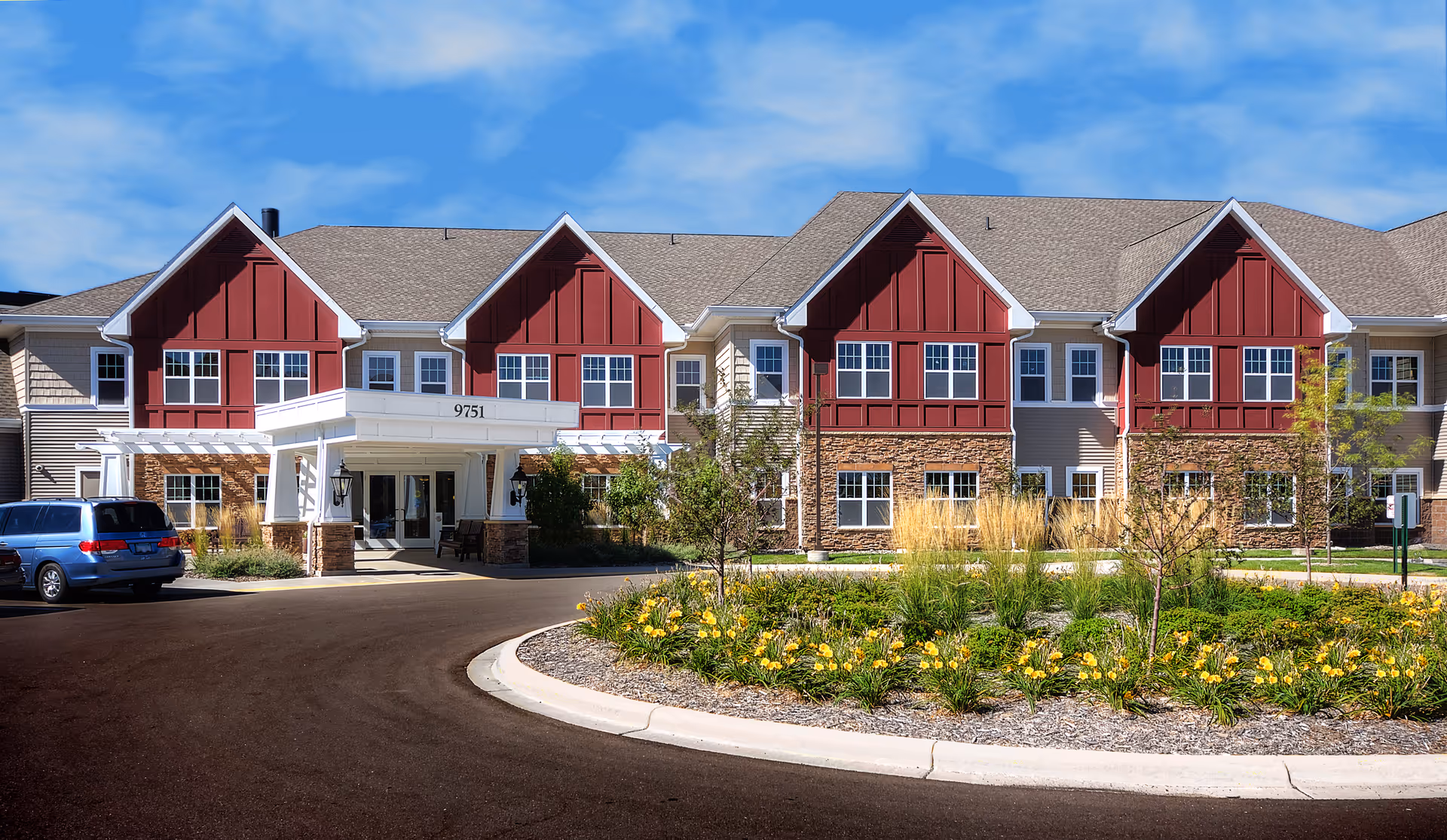 Front exterior view of a senior living facility building with beige and red siding, multiple windows, a covered entrance with the number 9751, a circular driveway, and landscaped flower beds with yellow flowers and greenery under a blue sky.