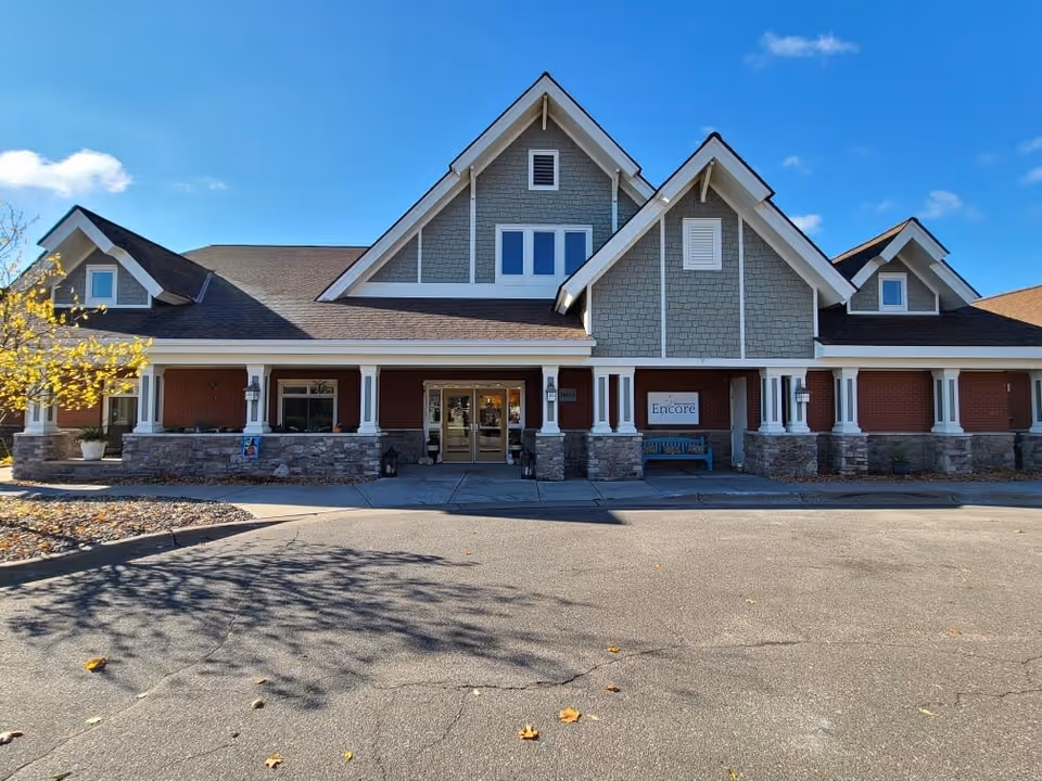 Front exterior view of Encore Assisted Living and Memory Care at North Branch building with a paved driveway, stone and brick facade, multiple gabled roofs, and a clear blue sky.