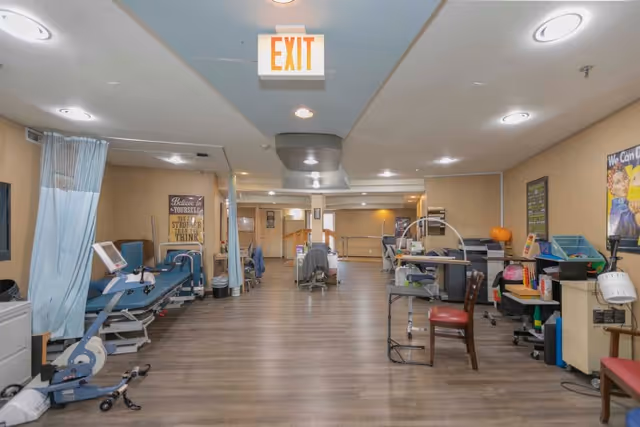 Interior view of a rehabilitation center room with exercise equipment, chairs, and medical devices arranged along the walls. The room has wood flooring, beige walls, and ceiling lights. There is an exit sign hanging from the ceiling and motivational posters on the walls.