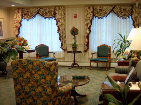 Cozy sitting room with floral-upholstered chairs, small tables, patterned drapes, and potted plants.
