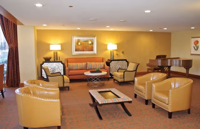 A cozy living room area with a sofa, four armchairs, two side tables with lamps, a coffee table, and a grand piano in the corner. The walls are painted yellow and decorated with framed artwork. There is a large window with curtains on the left side.