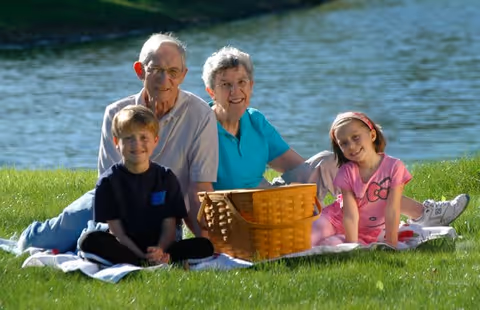 An elderly couple sitting on a blanket on the grass near a body of water, accompanied by a young boy and girl, with a picnic basket in front of them.