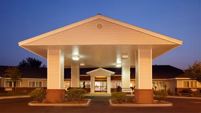 Front exterior view of Richland Assisted Living facility at dusk, showing a large covered entrance with pillars, landscaped bushes, and a well-lit building in the background.