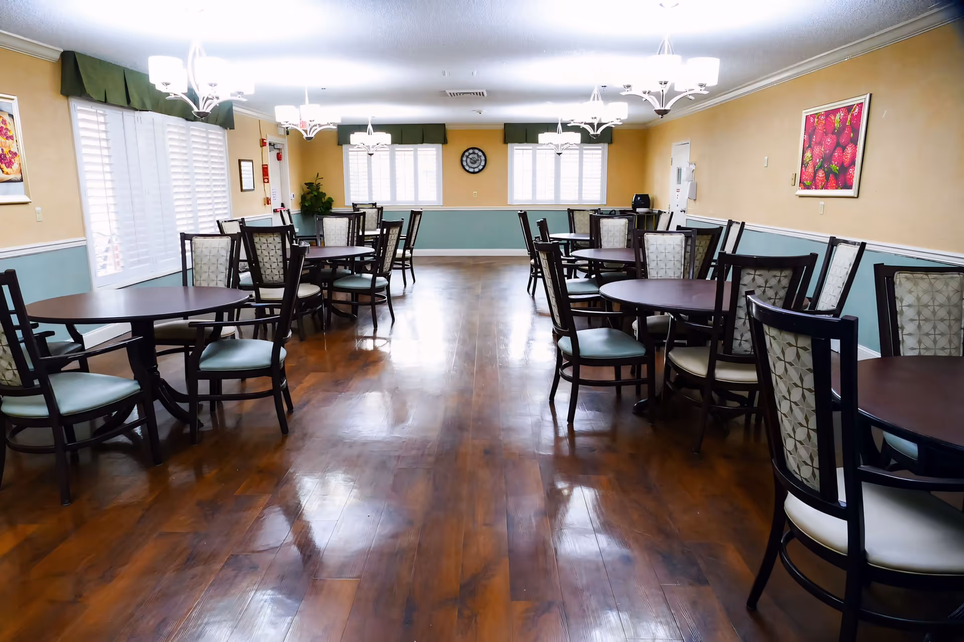 A spacious dining room with multiple round wooden tables and cushioned chairs arranged neatly. The room has wooden flooring, beige walls with a light blue wainscoting, and several windows with white shutters allowing natural light. There are chandeliers hanging from the ceiling and framed artwork on the walls.