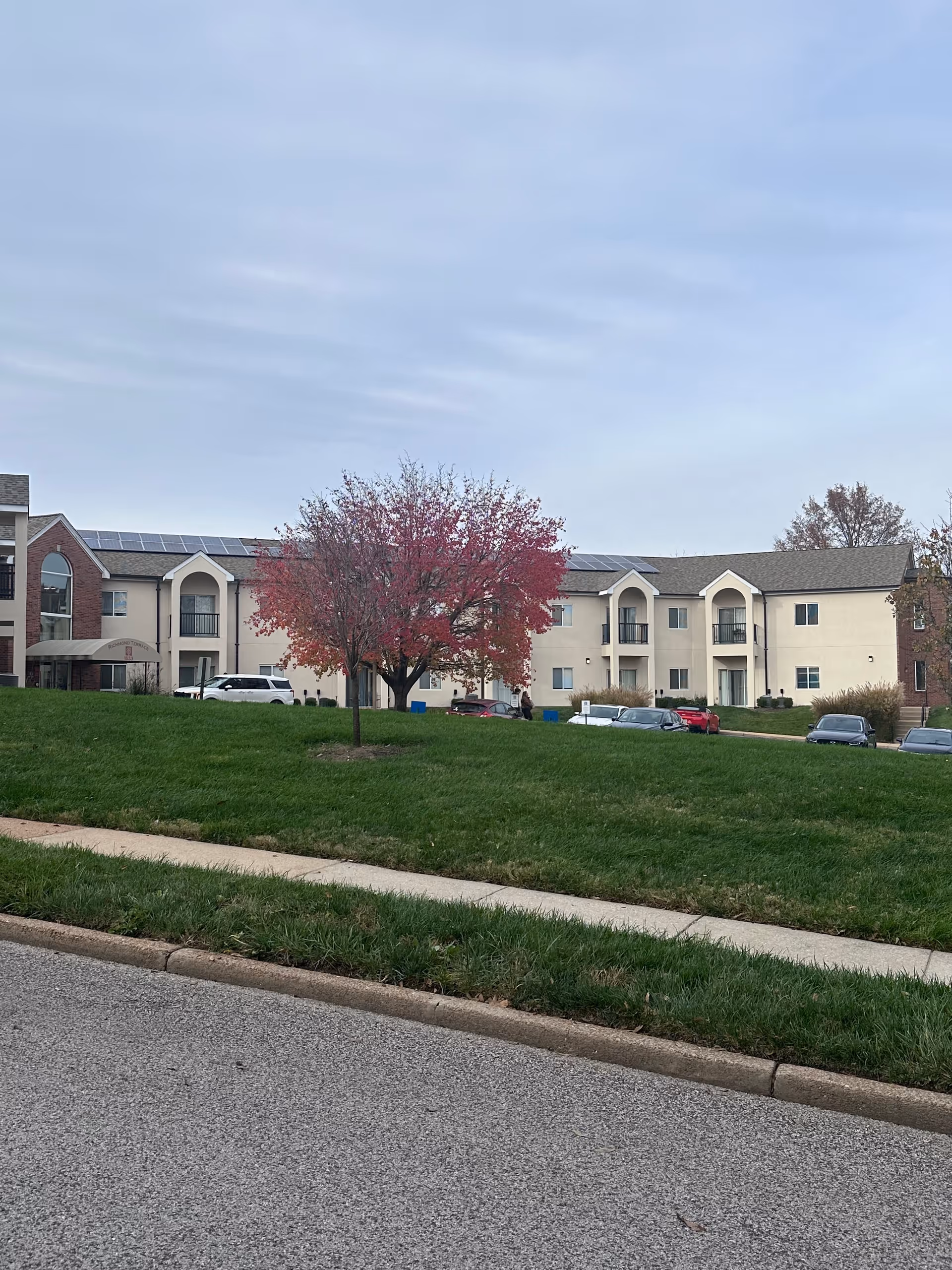 Exterior view of Richmond Terrace senior living facility showing a two-story building with beige and brick facade, solar panels on the roof, a tree with red leaves in front, several parked cars, and a grassy area with a sidewalk in the foreground under a cloudy sky.