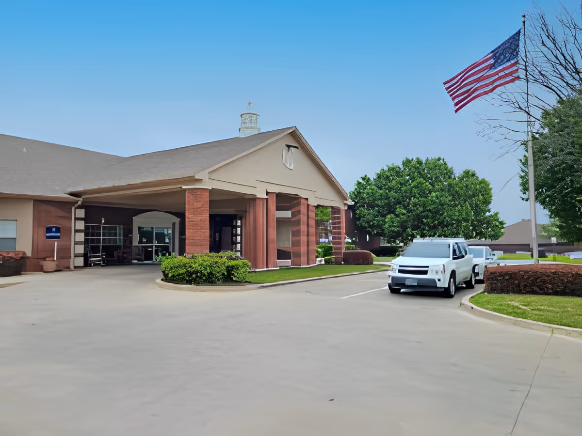 Exterior view of Morada Burleson facility showing the front entrance with a covered drop-off area, two parked white vehicles, an American flag on a flagpole, and surrounding greenery under a clear blue sky.