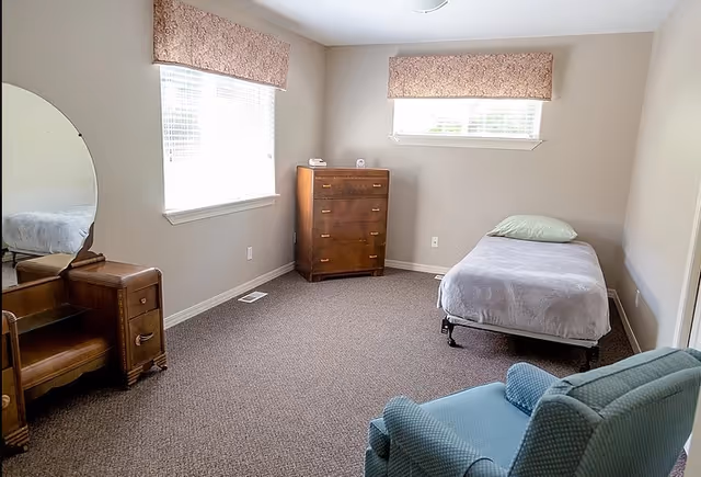 A simple bedroom in an assisted living facility with a single bed covered with a gray blanket and a green pillow, a wooden dresser against the wall, a wooden vanity with a mirror, and a blue upholstered armchair. The room has two windows with patterned valances and beige walls.