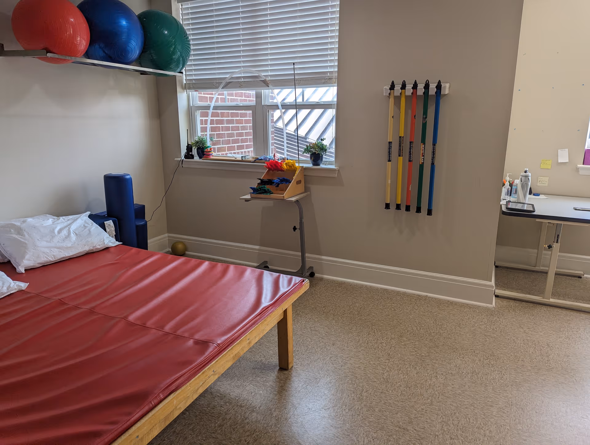 A therapy or exercise room with a padded red mat on a wooden frame, several large exercise balls on a shelf, colorful resistance bands hanging on the wall, a small table with therapy tools, and a window with blinds letting in natural light.
