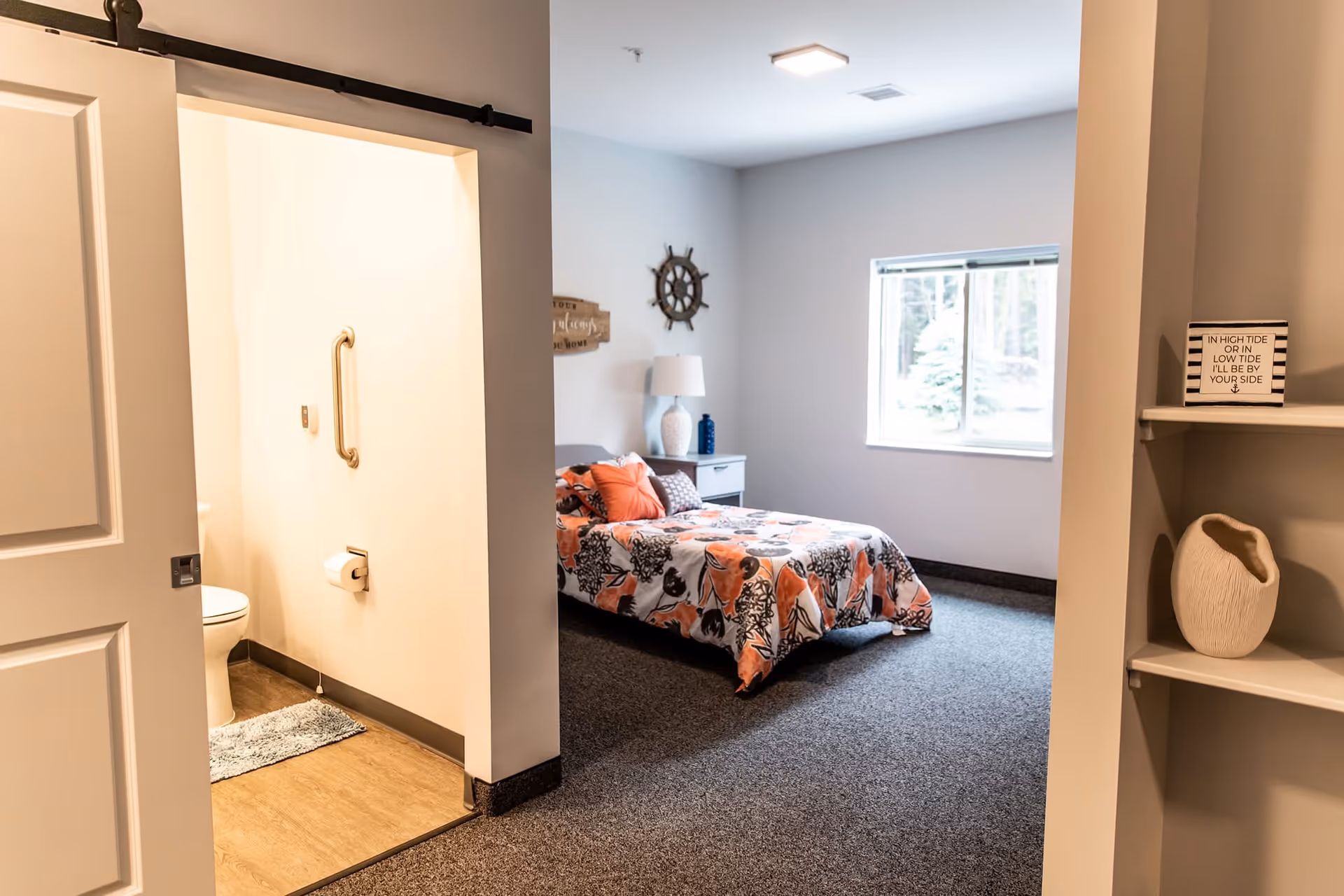 A cozy bedroom with a bed covered in a floral orange and black patterned comforter, a nightstand with a lamp and decorative items, a window letting in natural light, and an adjacent bathroom with a toilet and grab bar visible. A small shelf with a decorative vase and a sign that reads 'IN HIGH TIDE OR IN LOW TIDE I’LL BE BY YOUR SIDE' is also seen.