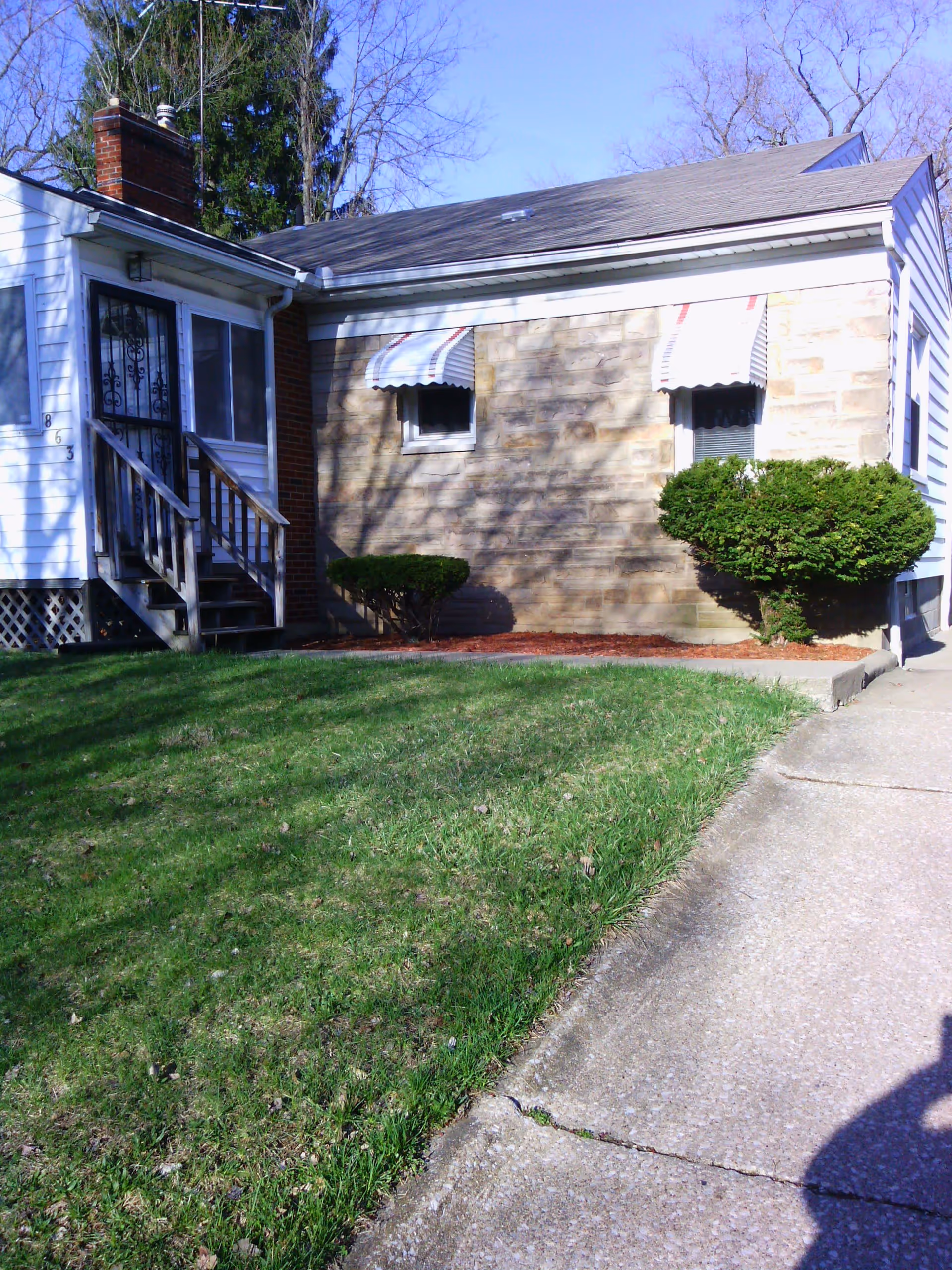 Exterior view of a single-story house with a brick chimney, two small windows with striped awnings, and a small porch with wooden steps. There is a green lawn in front and a concrete driveway on the right side. Some bushes are planted near the house.
