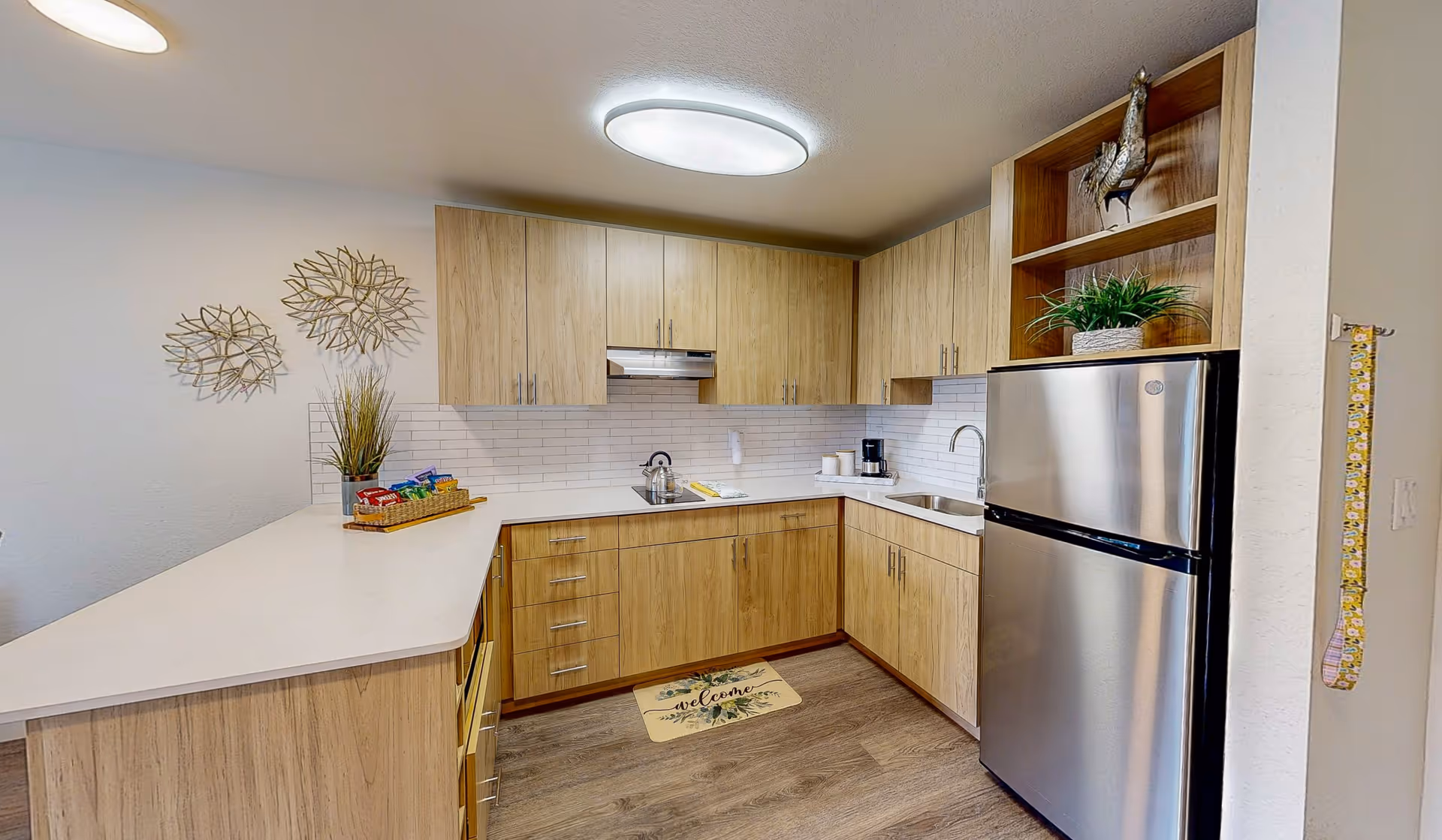 Modern kitchen with light wood cabinets, white countertops, a stainless steel refrigerator, a sink, a stove with a kettle, and decorative items including a plant and wall art. The floor is wood, and there is a welcome mat in front of the sink.