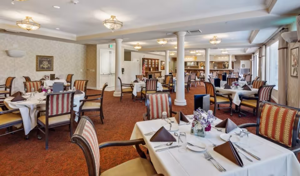 A spacious dining room with multiple tables set for a meal. Each table is covered with a white tablecloth and has brown folded napkins, plates, glasses, and silverware. The chairs have striped upholstery with wooden frames. The room features carpeted floors, decorative ceiling lights, and large windows with curtains allowing natural light to enter. There are white columns and a cabinet with an American flag in the background.