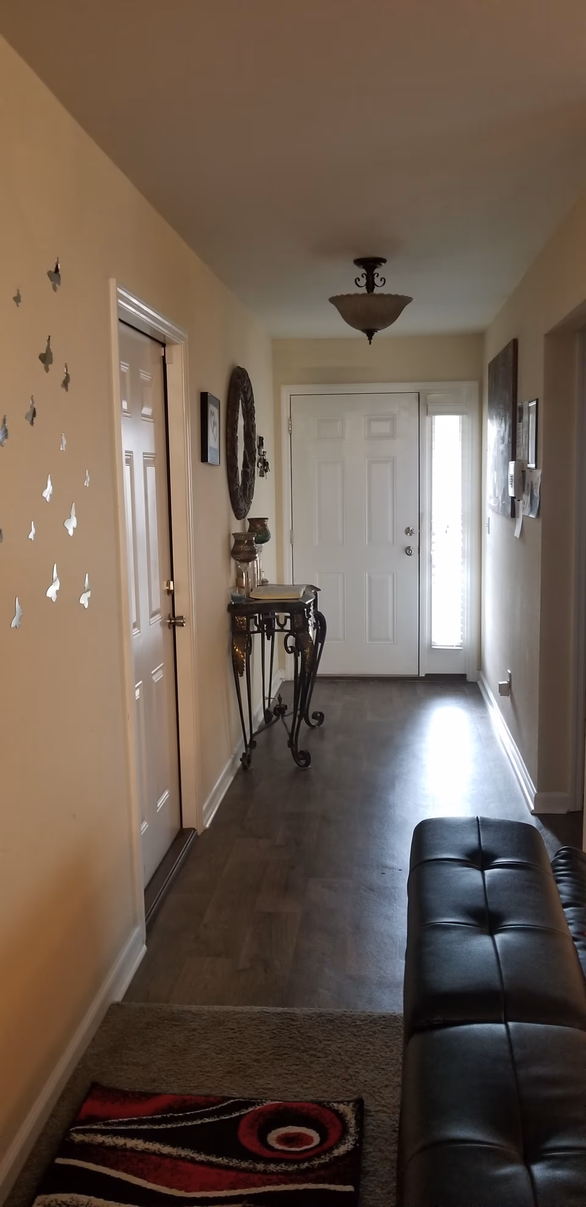 A hallway in an assisted living facility with a white front door at the end. The hallway has light-colored walls decorated with butterfly wall decals on the left side and framed pictures on the right. There is a decorative console table with a mirror above it along the left wall. A black leather bench is positioned on the right side near the foreground, and a small rug with red, black, and white patterns is on the floor.