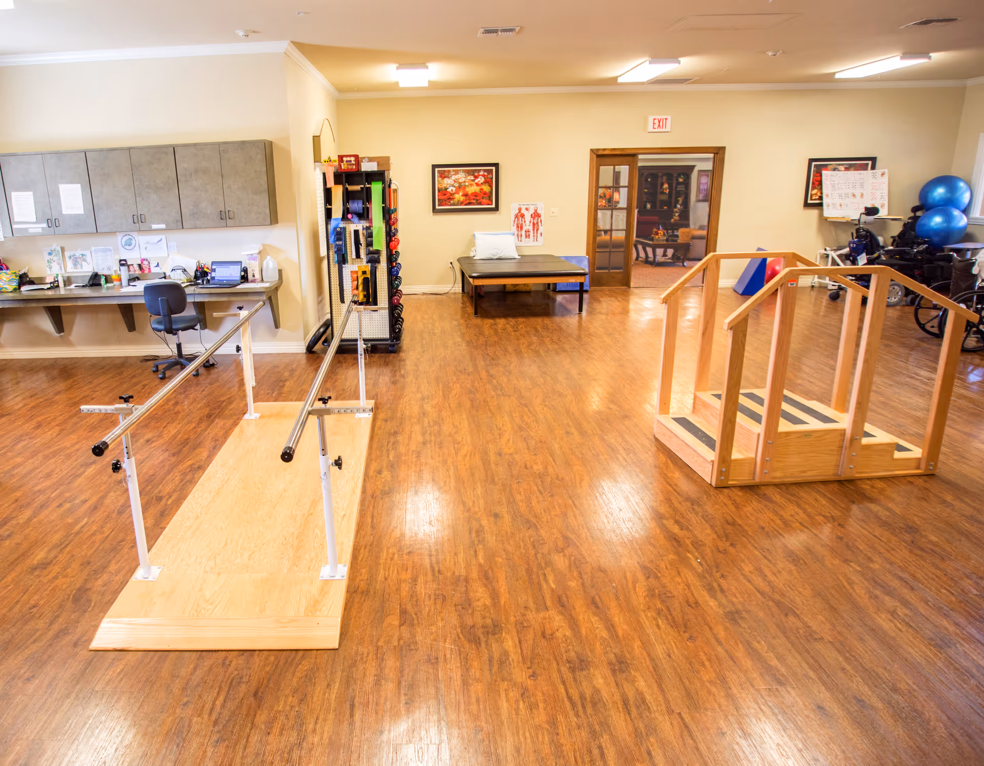A rehabilitation therapy room with wooden flooring, parallel bars for walking exercises, a small wooden staircase with handrails, exercise balls, a therapy table, and a desk with a chair and cabinets along the wall.
