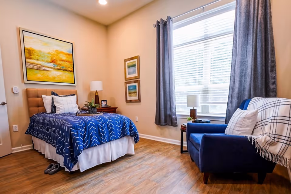 A cozy bedroom with a single bed covered in a blue patterned blanket and white pillows. There is a wooden nightstand with a lamp, a small plant, and a framed photo. The room has light-colored walls with three framed artworks, a large window with gray curtains, and a blue armchair draped with a white and black checkered throw. The floor is wooden, and a pair of slippers is placed near the bed.