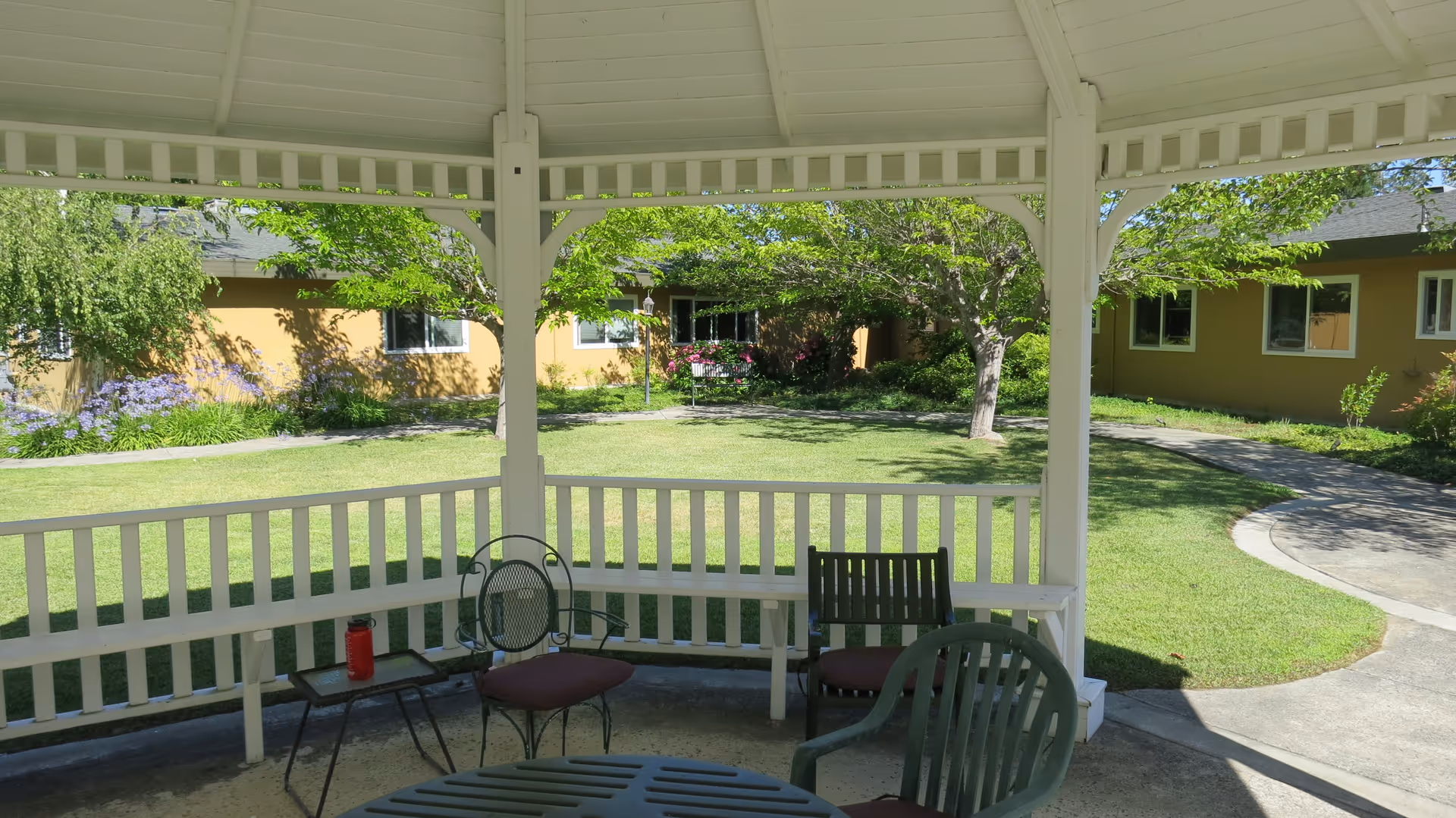 View from inside a white gazebo looking out onto a green lawn with trees and a yellow building in the background. The gazebo has a white railing and several chairs with cushions around a table. A red water bottle is on a small side table.