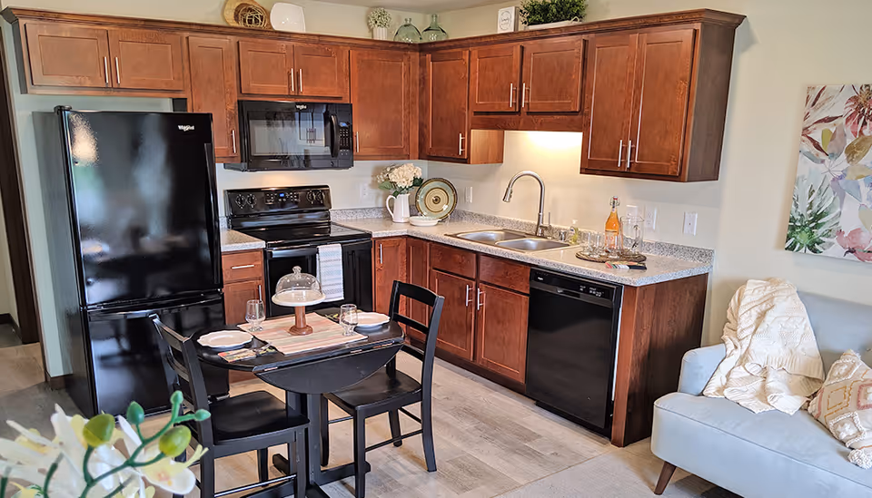 A modern kitchen area with dark wooden cabinets, black appliances including a refrigerator, microwave, stove, and dishwasher. A small dining table with two chairs is set with plates, glasses, and a cake stand. To the right, part of a light blue sofa with cushions and a throw blanket is visible, along with a colorful floral wall art.