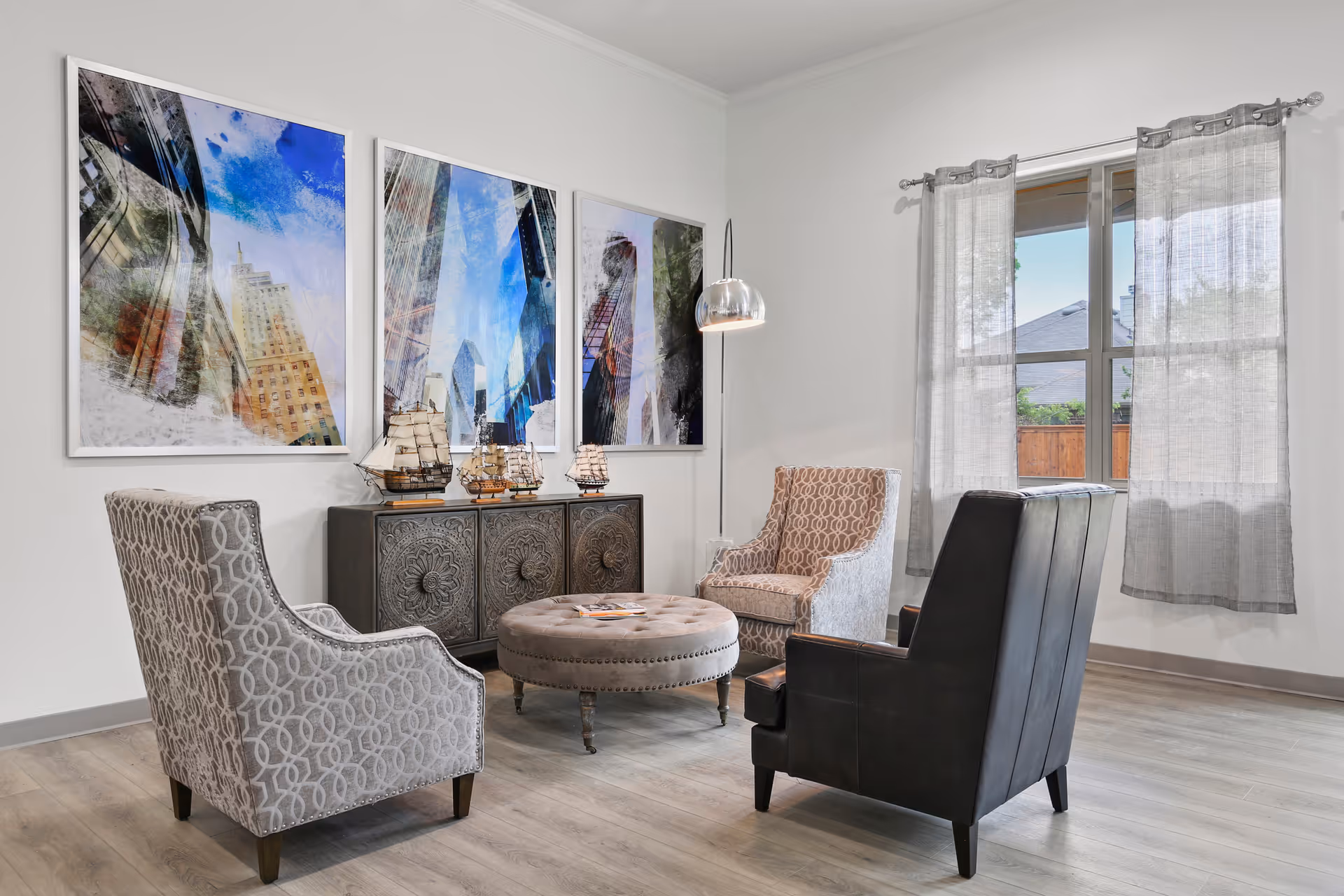 A cozy sitting area with three upholstered armchairs arranged around a round tufted ottoman. Behind the ottoman is a decorative cabinet with model ships on top. Three large abstract cityscape paintings hang on the wall above the cabinet. A tall floor lamp with a metallic shade stands next to one of the chairs. A window with light gray curtains lets in natural light, showing a glimpse of a wooden fence and roof outside.