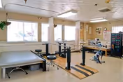 A physical therapy or rehabilitation room with exercise equipment including parallel bars for walking practice, a padded therapy table, stools, and various exercise machines. The room has large windows with curtains, light-colored walls, and a clean, organized appearance.
