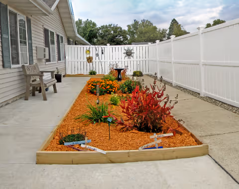 A small, neatly maintained garden bed with various plants and flowers, bordered by wooden planks and covered with mulch. The garden is situated between a beige building with windows and a white vinyl fence. There is a wooden bench on the left side near the building, and a concrete pathway runs alongside the garden bed.