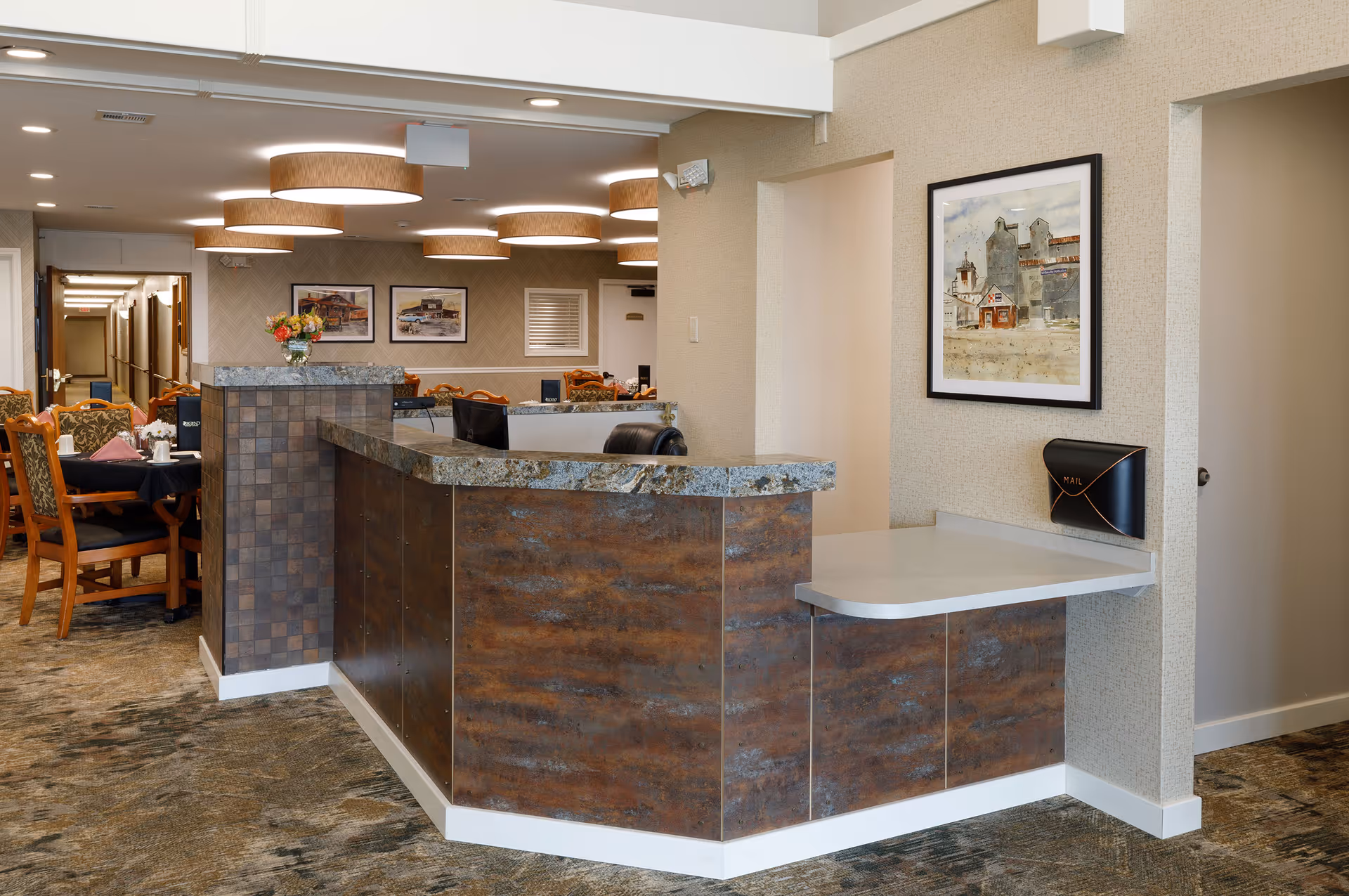 Reception desk area in Asbury Village senior living facility with a granite countertop and wood paneling. Behind the desk, there is a dining area with wooden chairs and tables set with napkins and cups. The walls are decorated with framed artwork and the ceiling has round, modern light fixtures. A small mail holder is mounted on the wall near the desk.