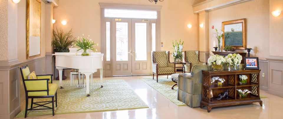 Well-lit reception lounge with seating, a white grand piano, floral arrangements, and double glass doors.