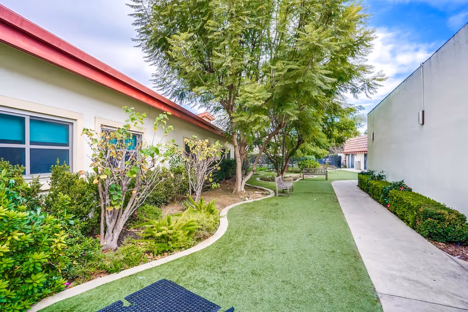 Outdoor garden area at Leisure Glen Care Center featuring a pathway with artificial grass, benches, trees, and bushes alongside a building with windows and a red roof edge under a partly cloudy sky.