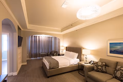 Spacious bedroom suite with a bed, nightstands and lamps, a sofa and armchair, and a large window with curtains.