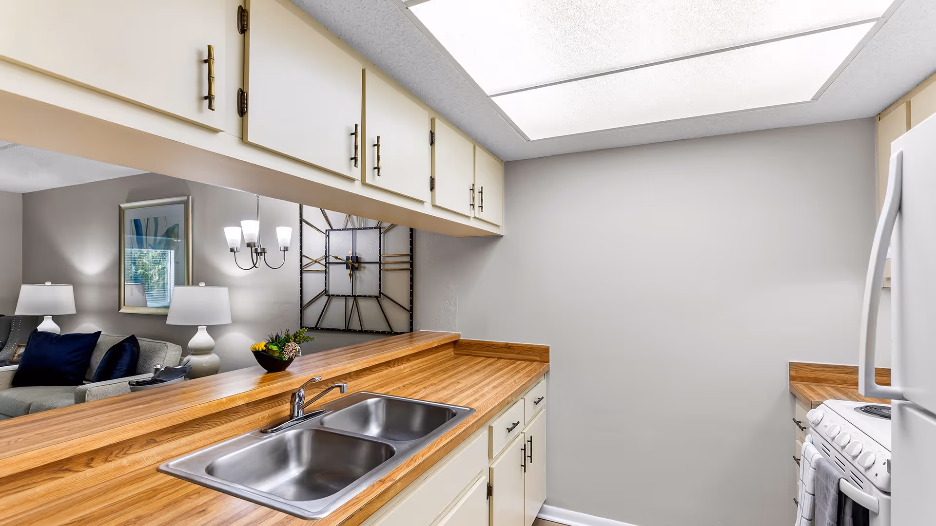 Interior view of a kitchen with wooden countertops, a double stainless steel sink, white cabinets with brass handles, and a white stove. The kitchen opens to a living room area with a gray couch, blue pillows, two white lamps, a large wall clock, and a chandelier. The walls are painted light gray.