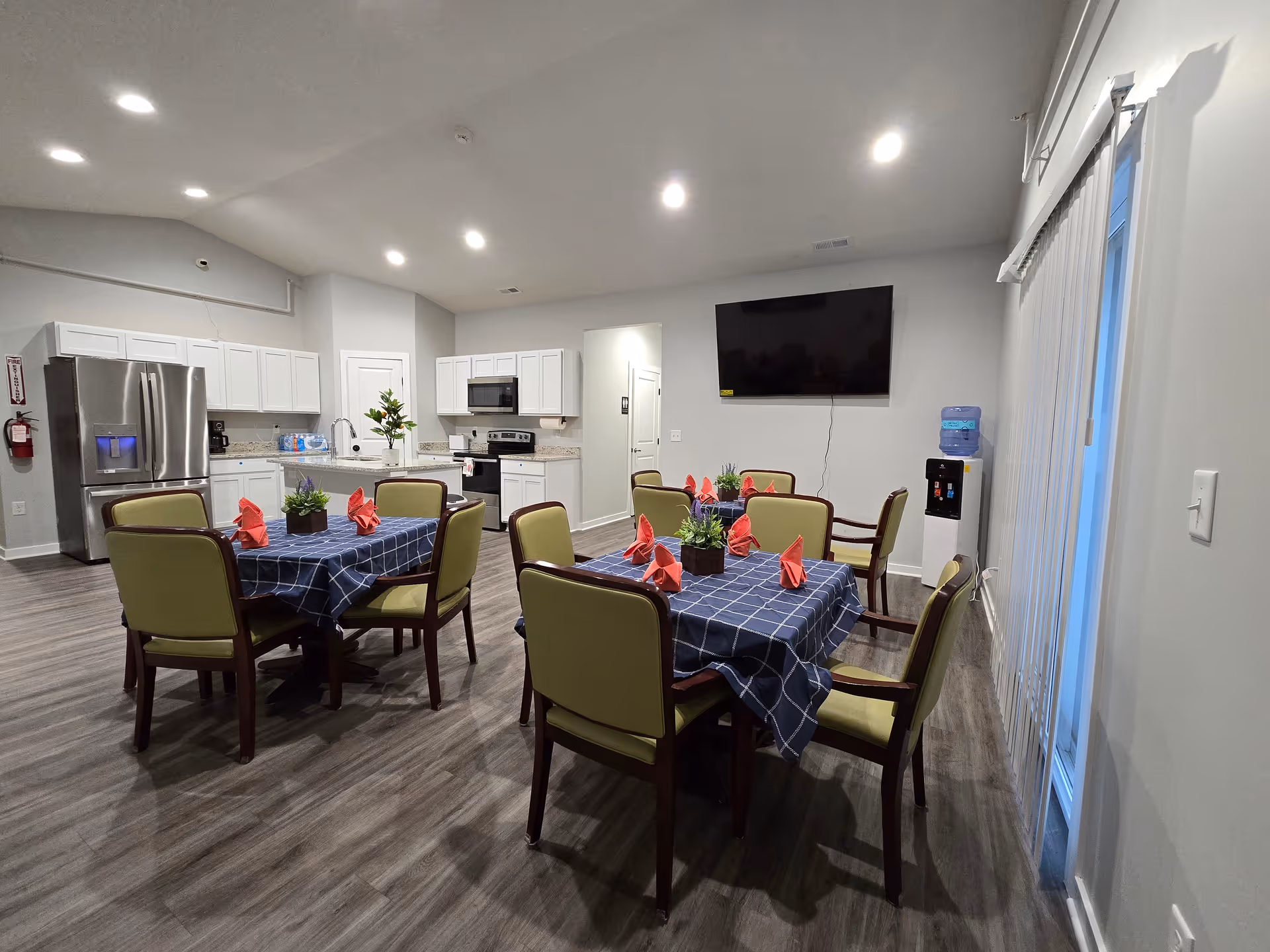 Open communal dining area with tables set with blue checkered tablecloths and red napkins, surrounding chairs, a kitchen with stainless appliances and a wall-mounted TV.
