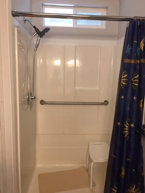 A bathroom shower area with a white plastic shower chair, a beige bath mat on the floor, a metal grab bar on the back wall, and a showerhead mounted on the left wall. There is a window above the shower area and a blue shower curtain with a celestial pattern partially drawn to the right.