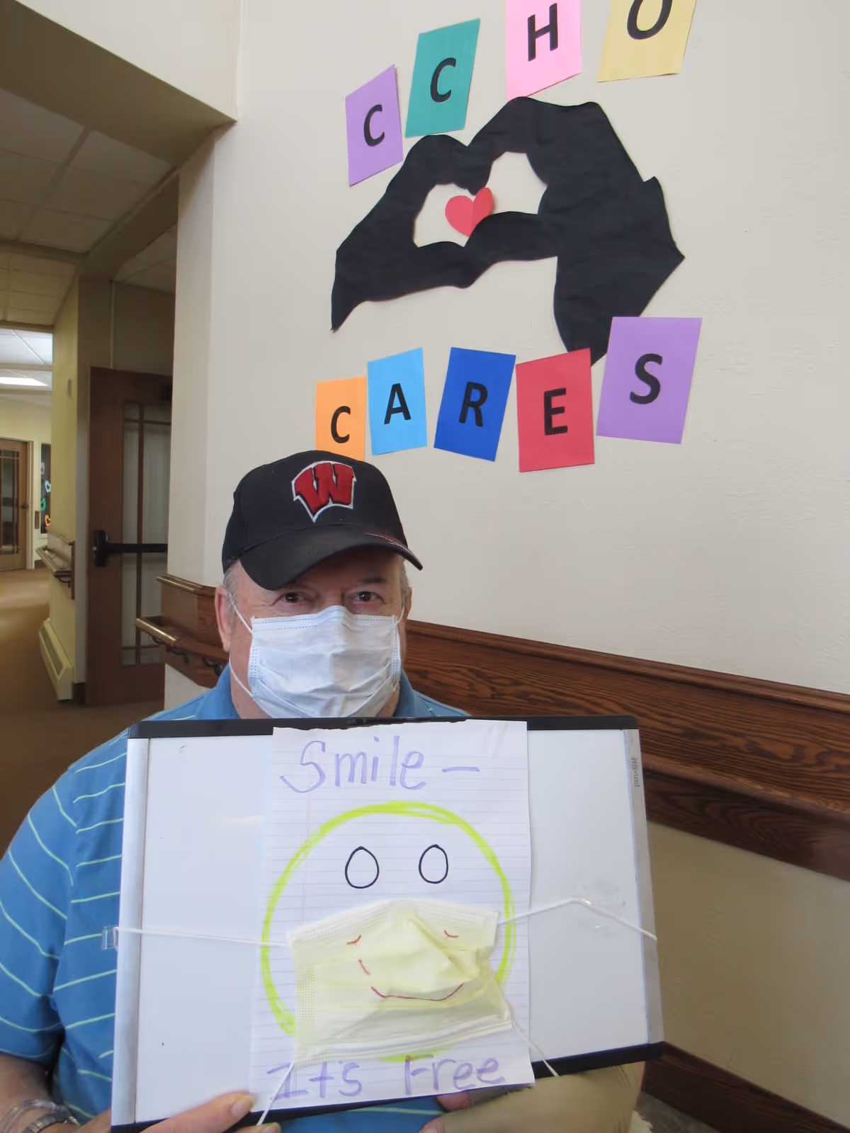 An elderly man wearing a face mask and a black cap with a red 'W' logo sits in a hallway. He holds a whiteboard with a paper attached that has a smiley face drawn on it, with a yellow face mask placed over the mouth area to resemble a smile. Behind him on the wall are colorful paper letters spelling out 'CCH CARES' with a heart shape made by two black hand cutouts.