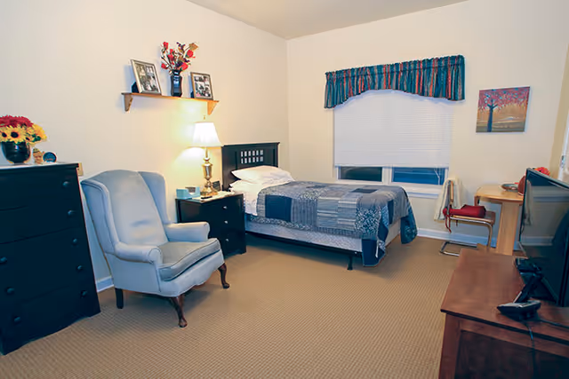 A cozy bedroom in a senior living facility featuring a single bed with a patchwork quilt, a light blue upholstered armchair, a wooden dresser with a vase of flowers, a nightstand with a lamp, framed photos on a wall shelf, a window with a colorful valance, a small desk with a chair, and a television on a wooden stand.