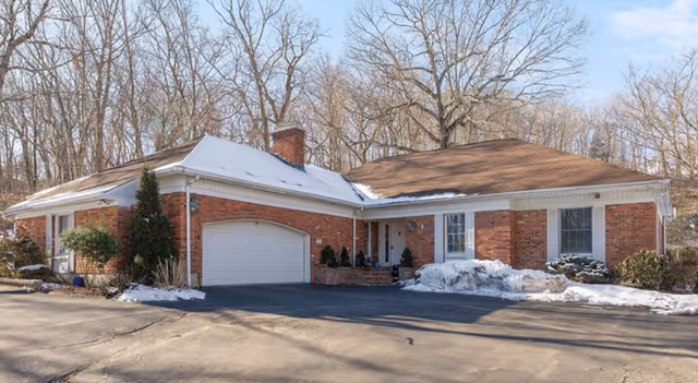 Front exterior of a single-story red brick ranch-style home with attached garage, driveway, and light snow on the roof and ground.