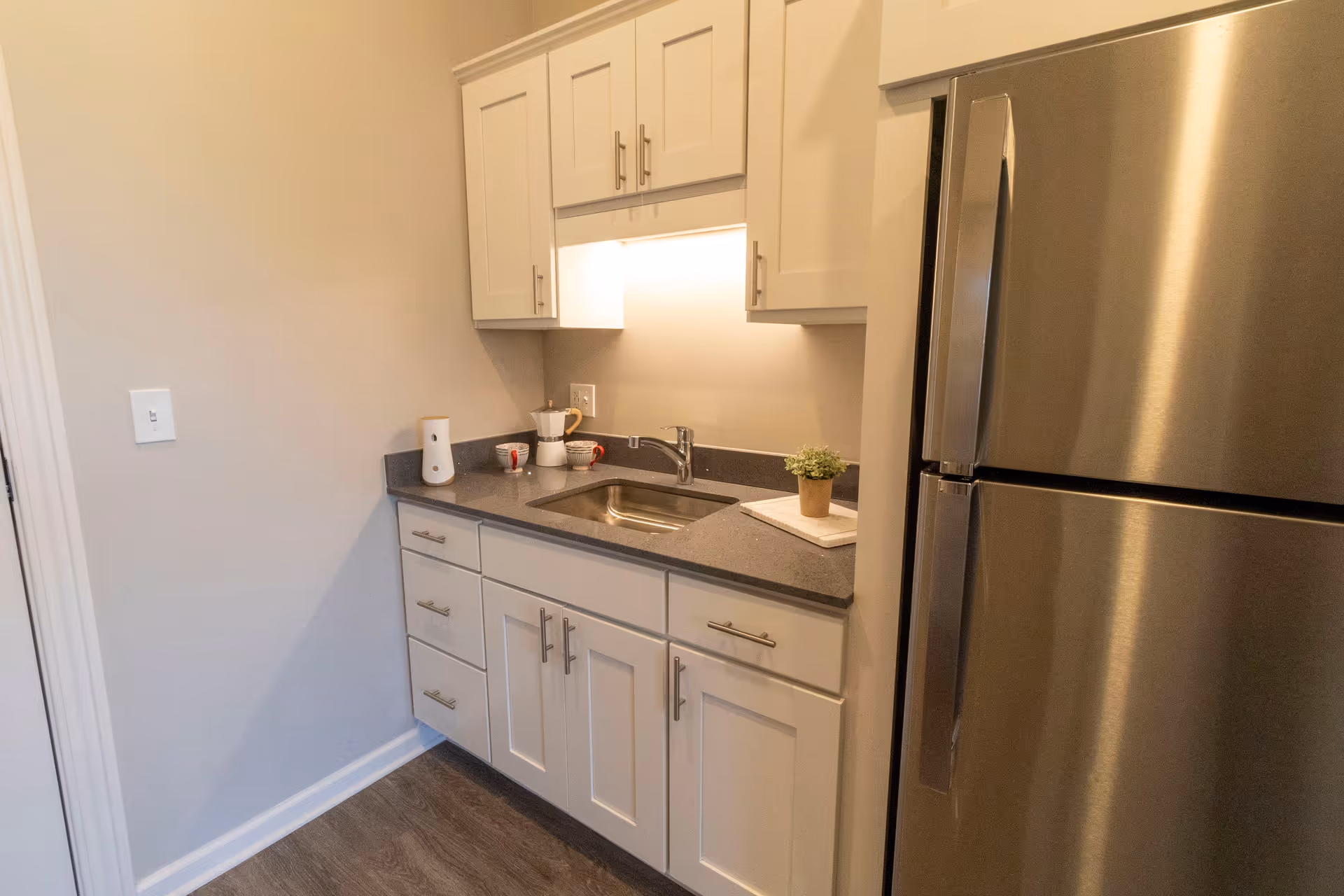Small kitchenette with white cabinets, a sink, countertop accessories and a stainless steel refrigerator.
