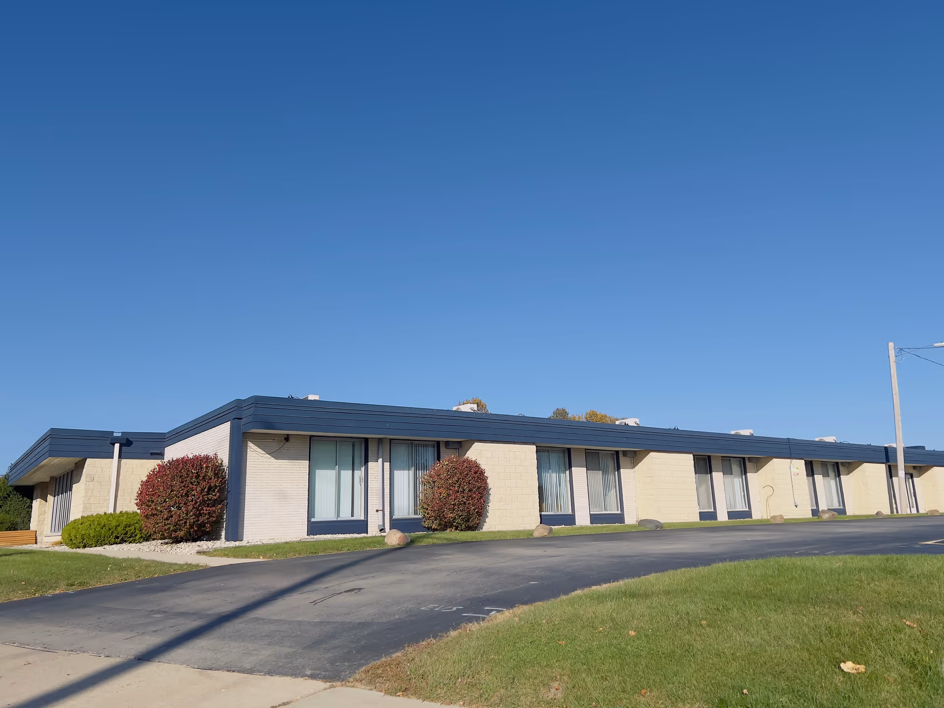 Single-story building with beige brick walls and a dark blue roof trim under a clear blue sky. The building has several large windows with vertical blinds and is surrounded by neatly trimmed bushes and a paved driveway.