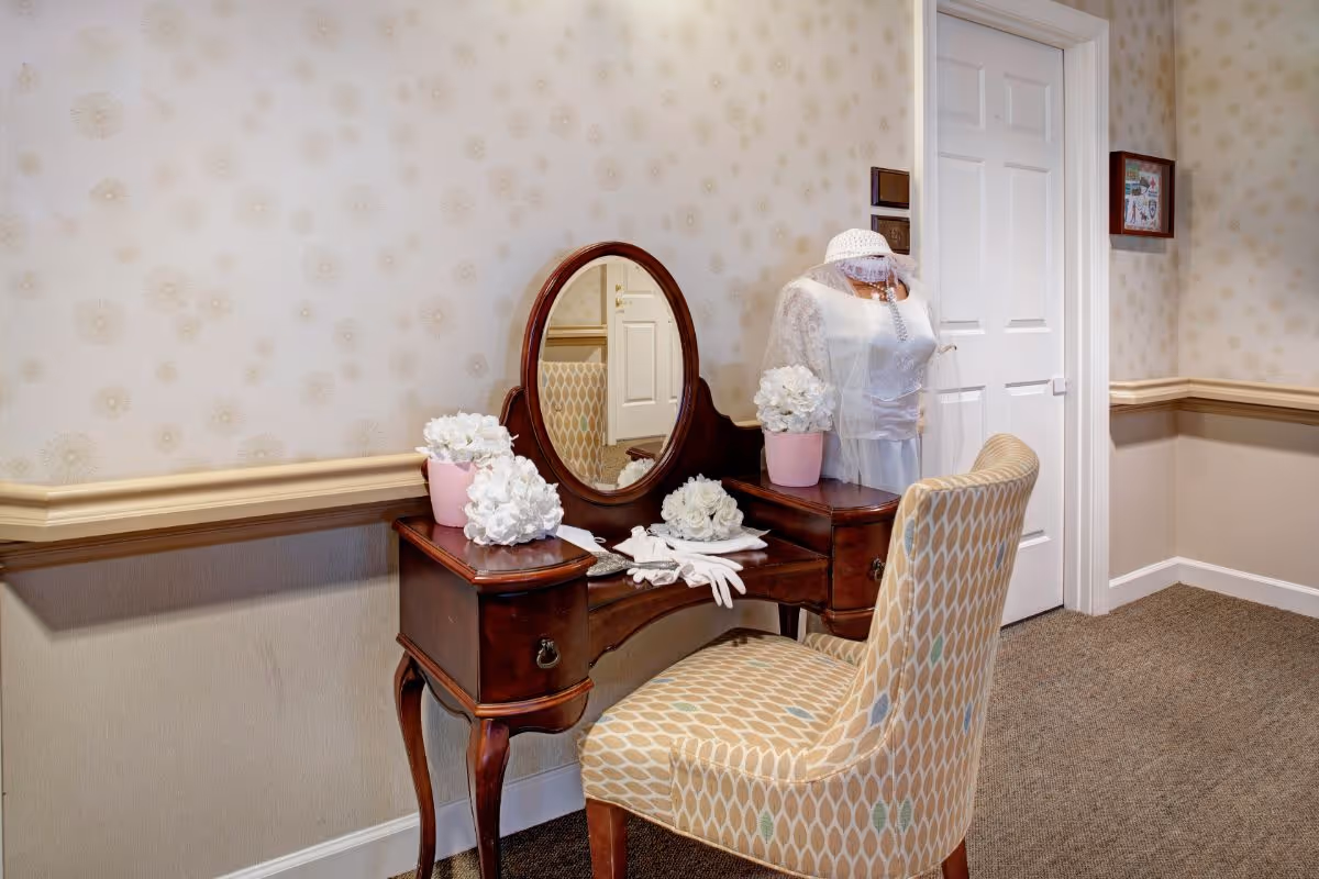 A vintage wooden vanity table with an oval mirror, decorated with white flowers in pink pots, white gloves, and a white lace bridal dress on a mannequin next to it. The setting is in a hallway with patterned wallpaper and a closed white door in the background.