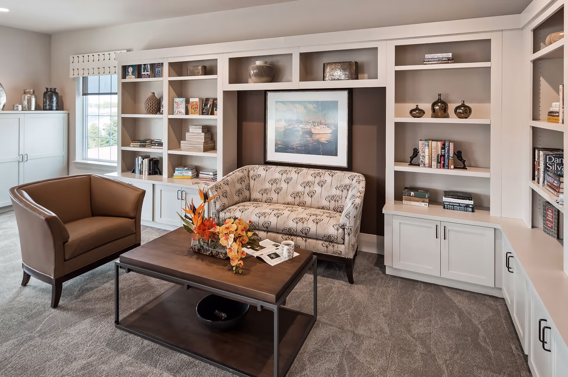 Cozy sitting area with a leather armchair, patterned loveseat, coffee table with flowers, and built-in bookshelves and cabinets.
