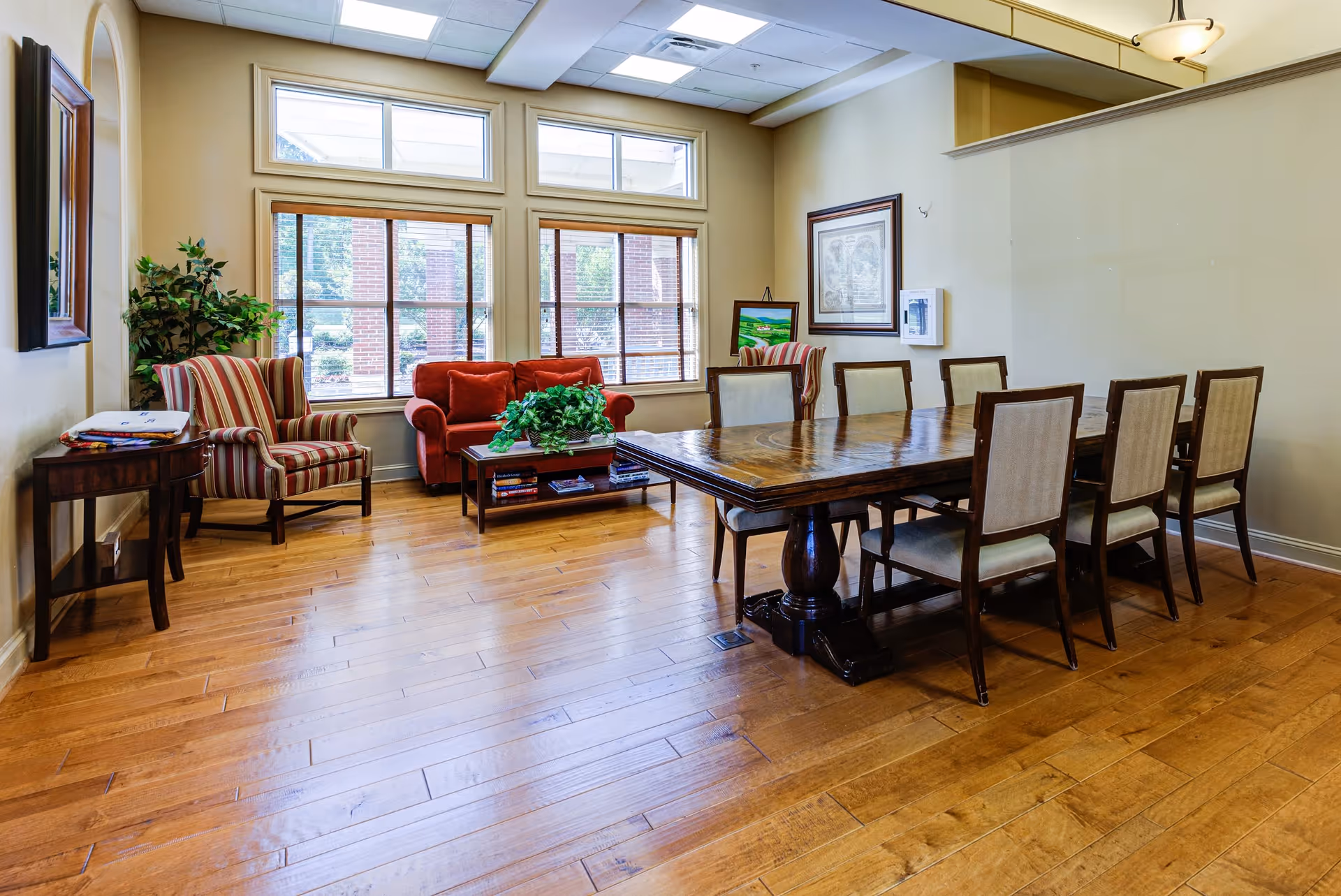 A well-lit room with a large wooden dining table surrounded by eight upholstered chairs. There is a red couch and a striped armchair near three large windows with blinds, allowing natural light to fill the space. The room has wooden flooring, a small side table with folded blankets, a potted plant, framed artwork on the walls, and a ceiling light fixture.