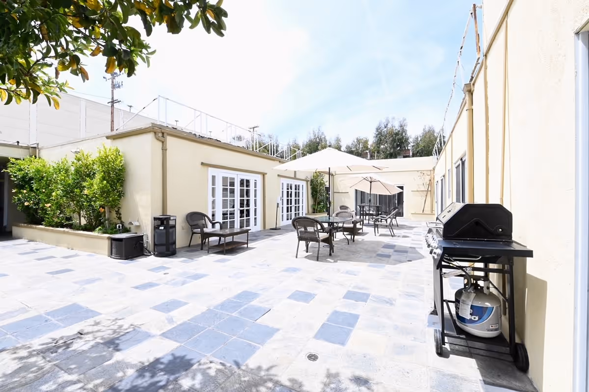 Sunny outdoor courtyard patio with tables, chairs, umbrellas, and a barbecue grill beside a low building.