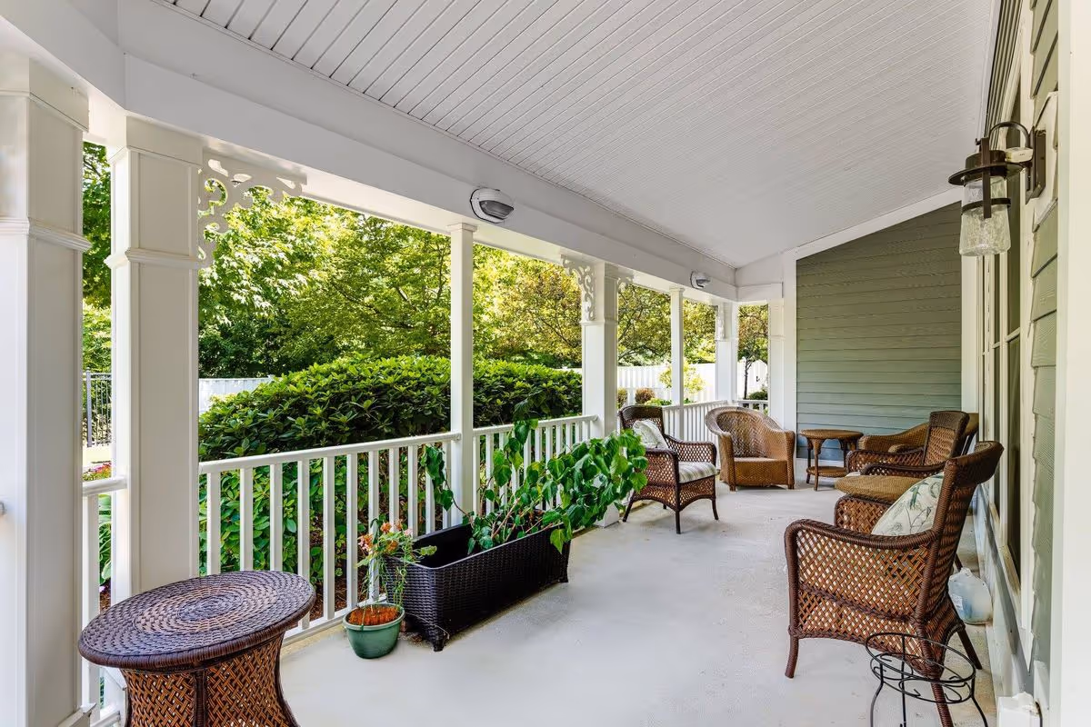 A covered outdoor porch area with white railings and ceiling, featuring several brown wicker chairs with cushions, a small wicker table, and potted plants. Green bushes and trees are visible beyond the porch.