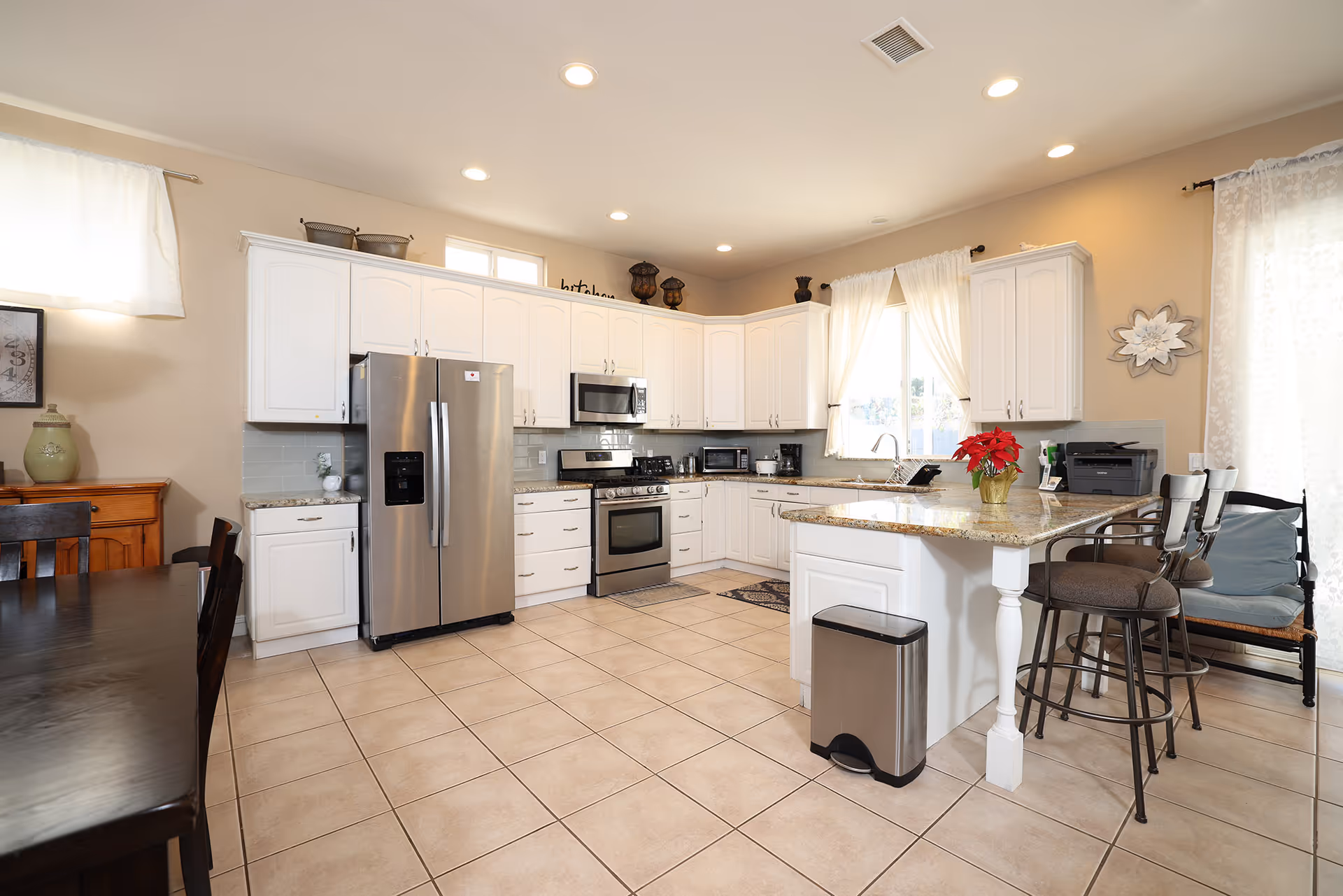 Bright and spacious kitchen with white cabinets, stainless steel refrigerator, stove, microwave, and granite countertops. There is a kitchen island with two bar stools, a trash can, and a small red poinsettia plant on the island. The room has tiled floors, beige walls, and windows with white curtains allowing natural light to enter.