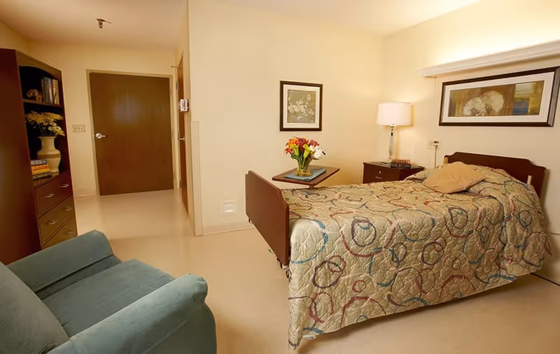 A furnished nursing facility bedroom with a single bed, bedside table and lamp, armchair, and a door to the hallway.
