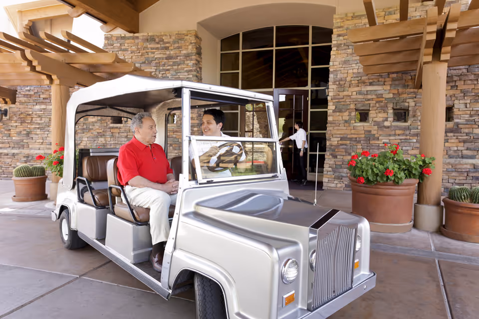 An elderly man in a red shirt sitting in a silver golf cart with a younger man driving. They are outside a building with stone walls and large potted plants with red flowers. A person is opening the glass door of the building in the background.