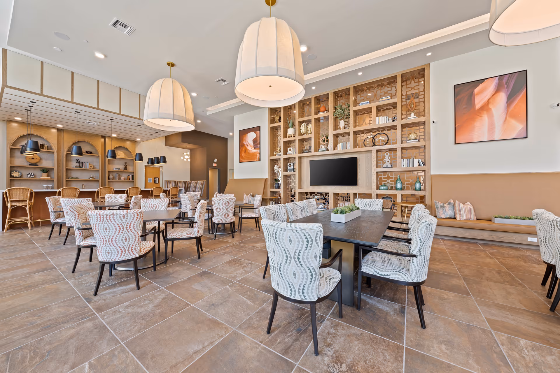 A spacious dining room with multiple tables and patterned chairs arranged neatly. The room features large pendant lights hanging from the ceiling, a built-in wooden shelving unit with decorative items and a flat-screen TV, and a long bench with cushions along one wall. The floor is tiled with large brown tiles, and there are framed abstract artworks on the walls.