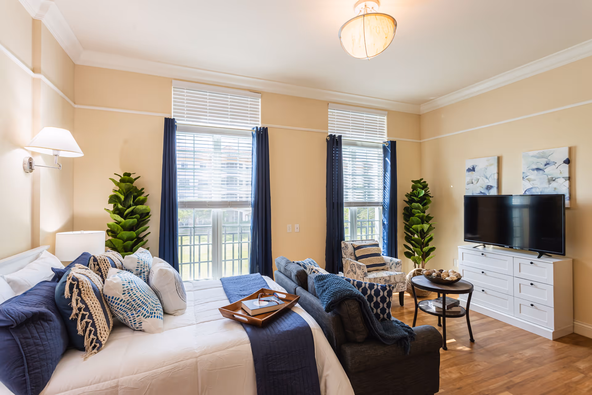 Bright, well-decorated bedroom suite with a made bed, seating area, TV on a dresser, and two tall windows with blue curtains.
