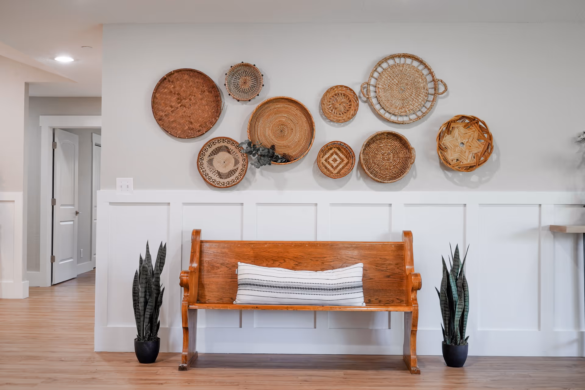 A wooden bench with a white and black striped pillow is placed against a white paneled wall. Above the bench, there is a decorative arrangement of various woven baskets in different sizes and patterns. Two potted snake plants are positioned on either side of the bench. The floor is light wood, and an open doorway is visible to the left.